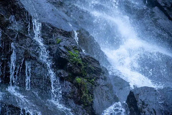 stock image A smiling man stands near a cascading waterfall on wet, dark rocks, wearing a backpack and outdoor gear, enjoying a nature hike