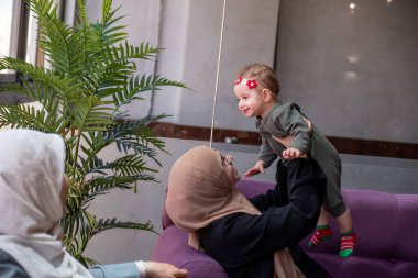 İki kadın tesettürlü ve bir çocuk bir kafede oturup sohbet ediyor ve kahvenin tadını çıkarıyor. Sahne sıcak, aile dostu bir atmosfer ve günlük sosyal etkileşimi tasvir ediyor..