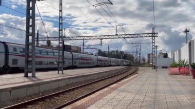 Fas 'taki modern yüksek hızlı Al Boraq TGV treni bulutlu bir günde ıssız bir istasyon platformuna ulaşarak, gelişmiş ulaşım altyapısı ve sakin çevresinin görüntülerini sergiliyor..