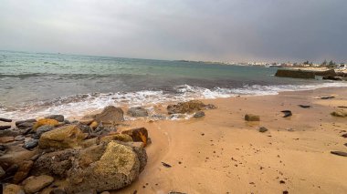 Okyanus dalgaları kumlu bir sahilde kıyıya vuruyor ön planda kayalar ve bulutlu bir gökyüzünün altında uzak bir kasaba.