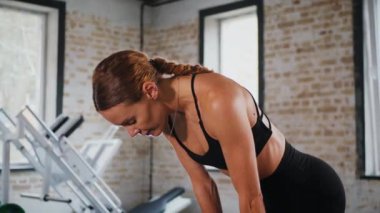 Atletik giyinmiş genç bir kadın parlak bir spor salonunda dikiliyor. Spor salonunda kararlı görünen bir kadın. Spor salonundaki antrenmana ara veren zinde bir kadın. Spor salonunda spor aletlerinin önünde poz veren kendine güvenen bir kadın.. 