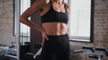 Atletik giyinmiş genç bir kadın parlak bir spor salonunda dikiliyor. Spor salonunda kararlı görünen bir kadın. Spor salonundaki antrenmana ara veren zinde bir kadın. Spor salonunda spor aletlerinin önünde poz veren kendine güvenen bir kadın.. 