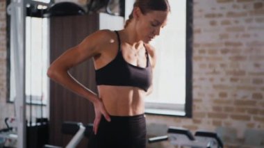 Atletik giyinmiş genç bir kadın parlak bir spor salonunda dikiliyor. Spor salonunda kararlı görünen bir kadın. Spor salonundaki antrenmana ara veren zinde bir kadın. Spor salonunda spor aletlerinin önünde poz veren kendine güvenen bir kadın.. 