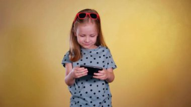 Güneş gözlüğü takan küçük bir kız cep telefonuyla oyun oynuyor. Güneş gözlüklü genç bir kız cep telefonuna bakıyor ve mobil bir sanal oyun oynarken mutlu bir şekilde gülüyor. Çocuk seviniyor, cep telefonu tutuyor. Stüdyoda sarı arka plan.