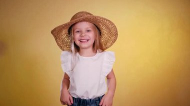 Hasır şapkalı gülümseyen küçük kız neşeyle dans eder ve güneşli sarı arka planda poz verir. Parlak sarı arka planda, güneş şapkalı mutlu çocuk. Sarı duvara hasır şapka takan şirin küçük bir kız. Hasır şapkalı sevimli kız. 