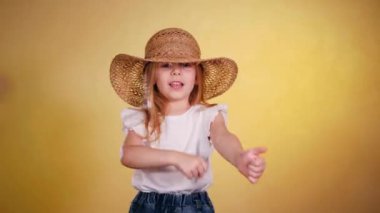 Hasır şapkalı gülümseyen küçük kız neşeyle dans eder ve güneşli sarı arka planda poz verir. Parlak sarı arka planda, güneş şapkalı mutlu çocuk. Sarı duvara hasır şapka takan şirin küçük bir kız. Hasır şapkalı sevimli kız. 