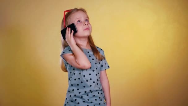 小さな女の子が携帯で幸せそうに話しかけている 携帯電話でチャットする若い女の子 笑顔の子供が携帯電話で会話をしています ジョイフルな子供が電話に出ました スタジオの黄色い背景 — ストック動画