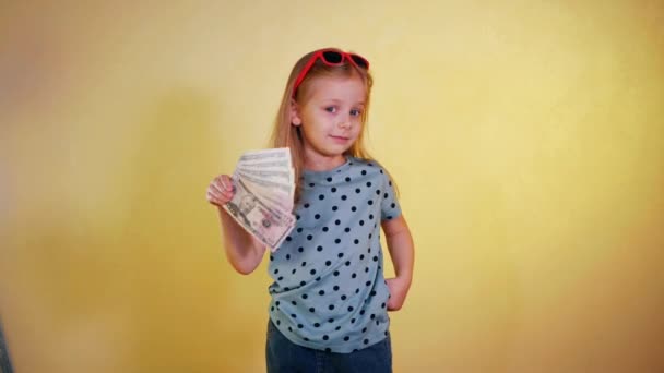 一个小女孩高兴地拿着一把现金 笑的孩子骄傲地炫耀着一堆钱 兴奋的女孩炫耀她的钱堆栈 带着一堆现金的快乐孩子小女孩挥金如土 黄色背景 — 图库视频影像