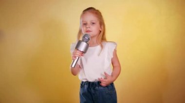 Genç bir kız parlak sarı bir arkaplan önünde kendine güvenen bir mikrofon tutuyor. Küçük bir şarkıcı kız, mikrofon tutuyor ve şarkı söylüyor. Çocuk sanatçı elinde mikrofonla sahneye çıkıyor, güneşli sarı bir renge karşı..