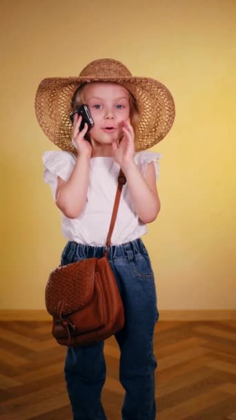 帽子をかぶったかわいい女の子が電話で笑う ストロー帽子の小さな女の子は 興奮して電話で話しています バーティカルビデオ 太陽の下の若い女の子は携帯電話でチャットします ストロー帽子の子供は 活発な電話会話を持っています かわいい — ストック動画