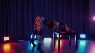 Dans eden seksi kadın twerk. Bir dansçı kız karanlık bir salonda erotik bir şekilde dans eder. Yavaş çekim. Twerk, yüksek topuklar, striptiz, baştan çıkarıcı bir dans. Atmosferik renkli ışık, kulüp, sahne. Balık ağı çoraplı bir kadın yerde boğucu bir poz veriyor..