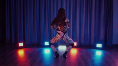 Dans eden seksi kadın twerk. Bir dansçı kız karanlık bir salonda erotik bir şekilde dans eder. Yavaş çekim. Twerk, yüksek topuklar, striptiz, baştan çıkarıcı bir dans. Atmosferik renkli ışık, kulüp, sahne. Balık ağı çoraplı bir kadın yerde boğucu bir poz veriyor..