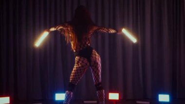 Elinde fenerlerle dans eden seksi bir kadın. Bir dansçı kız karanlık bir salonda erotik bir şekilde dans eder. Yavaş çekim. Twerk, yüksek topuklar, striptiz, baştan çıkarıcı bir dans. Balık ağı çoraplı bir kadın ahşap zeminde boğucu bir poz veriyor..