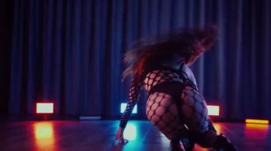 Dans eden seksi kadın twerk. Bir dansçı kız karanlık bir salonda erotik bir şekilde dans eder. Yavaş çekim. Twerk, yüksek topuklar, striptiz, baştan çıkarıcı bir dans. Atmosferik renkli ışık, kulüp, sahne. Balık ağı çoraplı bir kadın yerde boğucu bir poz veriyor..