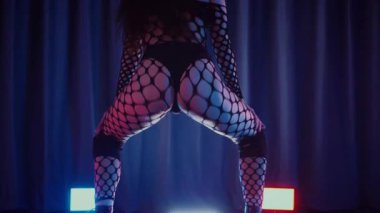 Dans eden seksi kadın twerk. Bir dansçı kız karanlık bir salonda erotik bir şekilde dans eder. Yavaş çekim. Twerk, yüksek topuklar, striptiz, baştan çıkarıcı bir dans. Atmosferik renkli ışık, kulüp, sahne. Balık ağı çoraplı bir kadın yerde boğucu bir poz veriyor..