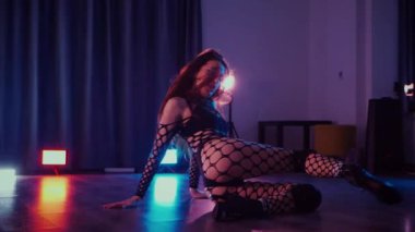Dans eden seksi kadın twerk. Bir dansçı kız karanlık bir salonda erotik bir şekilde dans eder. Yavaş çekim. Twerk, yüksek topuklar, striptiz, baştan çıkarıcı bir dans. Atmosferik renkli ışık, kulüp, sahne. Balık ağı çoraplı bir kadın yerde boğucu bir poz veriyor..