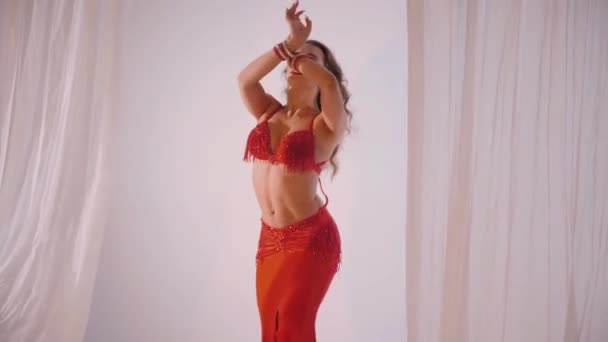 美丽的姑娘跳东方式肚皮舞 穿着红色内衣的性感女人正在一个白色工作室里跳着迷人的舞步 漂亮的肚子和乳房 穿着红色衣服的时髦女模特 穿着红色衣服摆姿势的优雅女人 — 图库视频影像