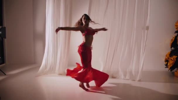 美丽的姑娘跳东方式肚皮舞 慢动作穿着红色内衣的性感女人正在一个白色工作室里跳着迷人的舞步 漂亮的肚子和乳房 穿着红色衣服的时髦女模特 穿着红色衣服摆姿势的优雅女人 — 图库视频影像