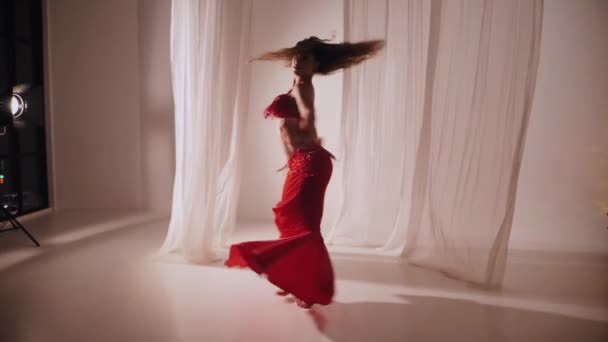 美丽的姑娘跳东方式肚皮舞 慢动作穿着红色内衣的性感女人正在一个白色工作室里跳着迷人的舞步 漂亮的肚子和乳房 穿着红色衣服的时髦女模特 穿着红色衣服摆姿势的优雅女人 — 图库视频影像