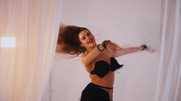 美丽的女孩跳东方式的肚皮舞 一个穿着黑色内衣的性感女人在一个白色的工作室里跳着迷人的舞步 漂亮的腹部和胸部 穿着黑色衣服的优雅女人优雅地跳舞 穿着黑色服装的迷人舞蹈家 — 图库视频影像