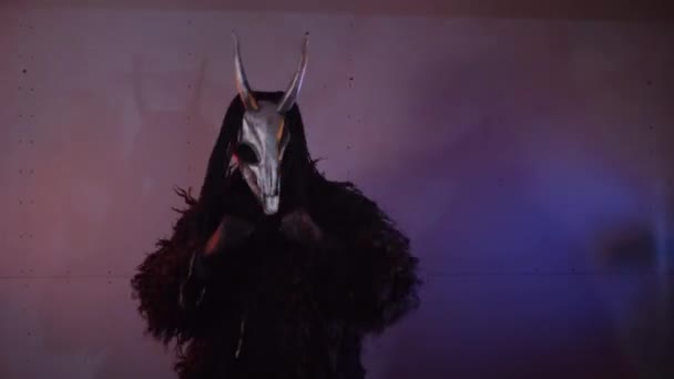 어두운 스튜디오에서 무서운 마스크를 신비한 남자는 검은색 코트와 머리에 뿔이있는 — 비디오