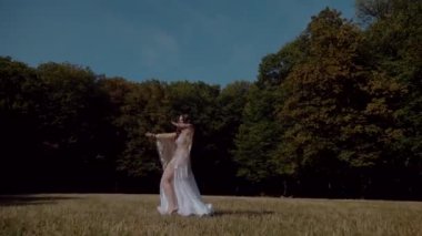 Ormanda göbek dansı yapan bir kız. Doğulu dansı. Beyaz göbek dansı kostümlü kadın sahada hareketlerle poz veriyor. Beyaz kostümlü zarif bir dansçı. Beyaz elbiseli kadın güzel bir açık havada göbek dansı hareketleri sergiliyor.