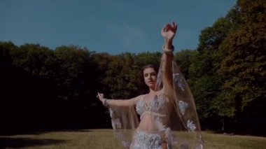 Ormanda göbek dansı yapan bir kız. Doğulu dansı. Beyaz göbek dansı kostümlü kadın sahada hareketlerle poz veriyor. Beyaz kostümlü zarif bir dansçı. Beyaz elbiseli kadın güzel bir açık havada göbek dansı hareketleri sergiliyor.