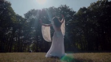 Ormanda göbek dansı yapan bir kız. Doğulu dansı. Beyaz göbek dansı kostümlü kadın sahada hareketlerle poz veriyor. Beyaz kostümlü zarif bir dansçı. Beyaz elbiseli kadın güzel bir açık havada göbek dansı hareketleri sergiliyor.