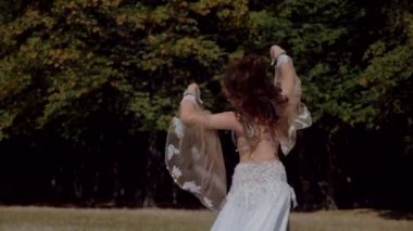 Ormanda göbek dansı yapan bir kız. Doğulu dansı. Ağır çekim. Sütyen ve etek içinde güzel bir dansçı. Beyaz göbek dansı kostümü giymiş bir kadın. Akan hareketlerle sahada poz veriyor. Beyaz giyinmiş bir kadın, güzel bir açık havada göbek dansı hareketleri sergiliyor.