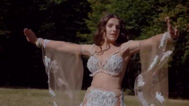 Ormanda göbek dansı yapan bir kız. Doğulu dansı. Ağır çekim. Sütyen ve etek içinde güzel bir dansçı. Beyaz göbek dansı kostümü giymiş bir kadın. Akan hareketlerle sahada poz veriyor. Beyaz giyinmiş bir kadın, güzel bir açık havada göbek dansı hareketleri sergiliyor.