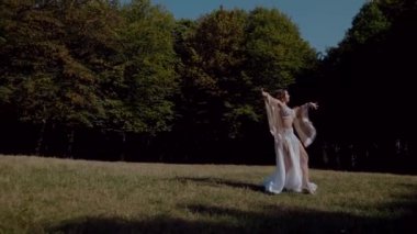 Ormanda göbek dansı yapan bir kız. Doğulu dansı. Ağır çekim. Sütyen ve etek içinde güzel bir dansçı. Beyaz göbek dansı kostümü giymiş bir kadın. Akan hareketlerle sahada poz veriyor. Beyaz giyinmiş bir kadın, güzel bir açık havada göbek dansı hareketleri sergiliyor.