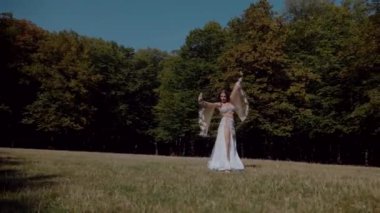 Ormanda göbek dansı yapan bir kız. Doğulu dansı. Ağır çekim. Sütyen ve etek içinde güzel bir dansçı. Beyaz göbek dansı kostümü giymiş bir kadın. Akan hareketlerle sahada poz veriyor. Beyaz giyinmiş bir kadın, güzel bir açık havada göbek dansı hareketleri sergiliyor.