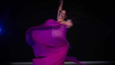 Mor elbiseli kadın loş ışıklı odada zarif bir şekilde göbek dansı yapıyor. Zarif giyimli kadın, müziğe gidiyor ve karanlık ortamda dans ediyor. Gölgeli alanda mor elbiseler giyen kadın dansçı. Karanlık odada dönen bir kadın. 
