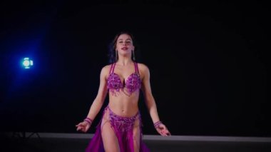 Mor elbiseli kadın loş ışıklı odada zarif bir şekilde göbek dansı yapıyor. Zarif giyimli kadın, müziğe gidiyor ve karanlık ortamda dans ediyor. Gölgeli alanda mor elbiseler giyen kadın dansçı. Karanlık odada dönen bir kadın. 