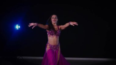 Mor elbiseli kadın loş ışıklı odada zarif bir şekilde göbek dansı yapıyor. Zarif giyimli kadın, müziğe gidiyor ve karanlık ortamda dans ediyor. Gölgeli alanda mor elbiseler giyen kadın dansçı. Karanlık odada dönen bir kadın. 