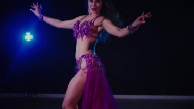 Mor elbiseli kadın loş ışıklı odada zarif bir şekilde göbek dansı yapıyor. Zarif giyimli kadın, müziğe gidiyor ve karanlık ortamda dans ediyor. Gölgeli alanda mor elbiseler giyen kadın dansçı. Karanlık odada dönen bir kadın. 