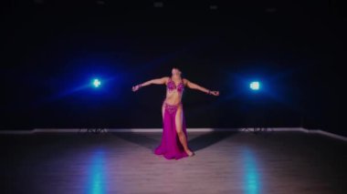 Mor elbiseli kadın loş ışıklı odada zarif bir şekilde göbek dansı yapıyor. Zarif giyimli kadın, müziğe gidiyor ve karanlık ortamda dans ediyor. Gölgeli alanda mor elbiseler giyen kadın dansçı. Karanlık odada dönen bir kadın. 