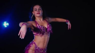 Mor elbiseli kadın loş ışıklı odada zarif bir şekilde göbek dansı yapıyor. Yavaş çekim. Zarif giyimli kadın, müziğe gidiyor ve karanlık ortamda dans ediyor. Mor elbiseler giyen kadın dansçı Gölgeli alanda girdaplar. 