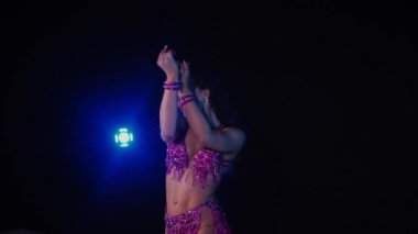 Mor elbiseli kadın loş ışıklı odada zarif bir şekilde göbek dansı yapıyor. Yavaş çekim. Zarif giyimli kadın, müziğe gidiyor ve karanlık ortamda dans ediyor. Mor elbiseler giyen kadın dansçı Gölgeli alanda girdaplar. 