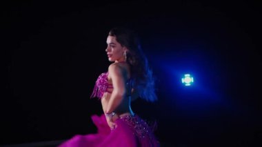 Mor elbiseli kadın loş ışıklı odada zarif bir şekilde göbek dansı yapıyor. Yavaş çekim. Zarif giyimli kadın, müziğe gidiyor ve karanlık ortamda dans ediyor. Mor elbiseler giyen kadın dansçı Gölgeli alanda girdaplar. 