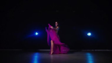 Mor elbiseli kadın loş ışıklı odada zarif bir şekilde göbek dansı yapıyor. Yavaş çekim. Zarif giyimli kadın, müziğe gidiyor ve karanlık ortamda dans ediyor. Mor elbiseler giyen kadın dansçı Gölgeli alanda girdaplar. 