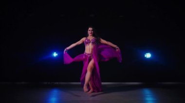 Mor elbiseli kadın loş ışıklı odada zarif bir şekilde göbek dansı yapıyor. Yavaş çekim. Zarif giyimli kadın, müziğe gidiyor ve karanlık ortamda dans ediyor. Mor elbiseler giyen kadın dansçı Gölgeli alanda girdaplar. 