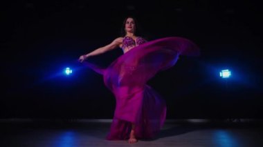 Mor elbiseli kadın loş ışıklı odada zarif bir şekilde göbek dansı yapıyor. Yavaş çekim. Zarif giyimli kadın, müziğe gidiyor ve karanlık ortamda dans ediyor. Mor elbiseler giyen kadın dansçı Gölgeli alanda girdaplar. 