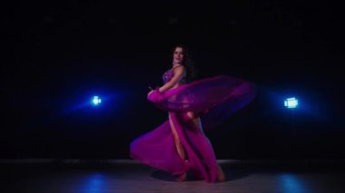 Mor elbiseli kadın loş ışıklı odada zarif bir şekilde göbek dansı yapıyor. Yavaş çekim. Zarif giyimli kadın, müziğe gidiyor ve karanlık ortamda dans ediyor. Mor elbiseler giyen kadın dansçı Gölgeli alanda girdaplar. 