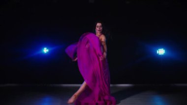 Mor elbiseli kadın loş ışıklı odada zarif bir şekilde göbek dansı yapıyor. Yavaş çekim. Zarif giyimli kadın, müziğe gidiyor ve karanlık ortamda dans ediyor. Mor elbiseler giyen kadın dansçı Gölgeli alanda girdaplar. 