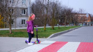 Küçük bir kız yaya geçidinden karşıya geçiyor. Kız bir scooter tutuyor ve yaya geçidi boyunca yolda yürüyordu. Yavaş çekim..