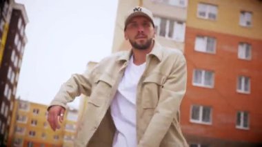 Şehir merkezinde hip hop dansı yapan bir adam. Adam havalı dans ediyor. Şehirdeki evlerin yakınında bir dansçı dans ediyor. Binanın dışında bej kıyafetli şık bir adam. Beyaz pantolonlu ve bej ceketli modaya uygun bir adam. Modaya uygun bir adam.