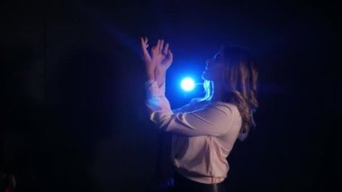 Karanlık bir odada poz veren genç bir kadın. Stüdyodaki seksi genç kız modeli. Bir kadın duygularını ve duygularını gösterir. Çekici kız kameraya bakıyor. Siyah arka plan, mavi spot ışığı, beyaz bluz..