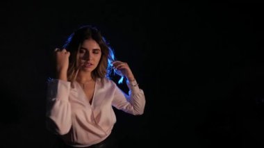 Karanlık bir odada poz veren genç bir kadın. Stüdyodaki seksi genç kız modeli. Bir kadın duygularını ve duygularını gösterir. Çekici kız kameraya bakıyor. Siyah arka plan, mavi spot ışığı, beyaz bluz..