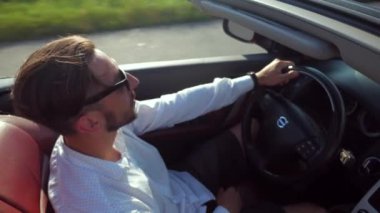 Adam üstü açık bir arabaya biniyor. Güzel kırmızı Cabriolet araba yüksek hızda yolda gidiyor. Seyahat rüyası. Güneşli bir günde güneş gözlüklü bir adam araba sürüyor. Trafikte sürücü güneş gözlüğü takıyor.. 